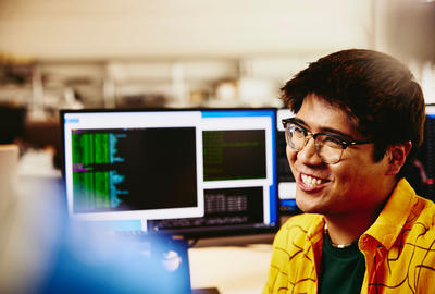 male smiling while sitting behind his desk, computer screens displaying programming code.