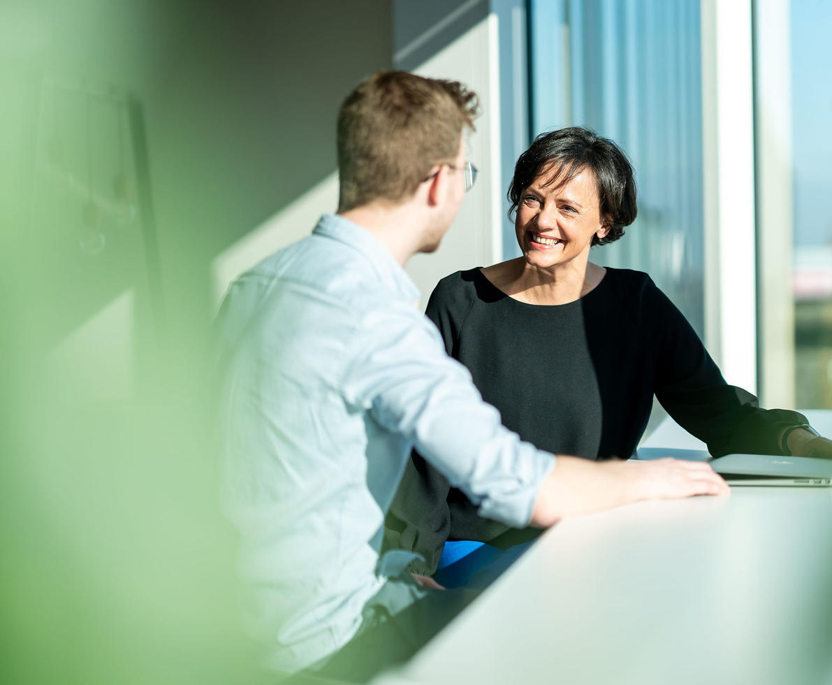 Photo of 2 colleagues talking and sitting at a table.